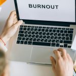 Overhead view of a person typing 'BURNOUT' on a laptop, surrounded by office materials.
